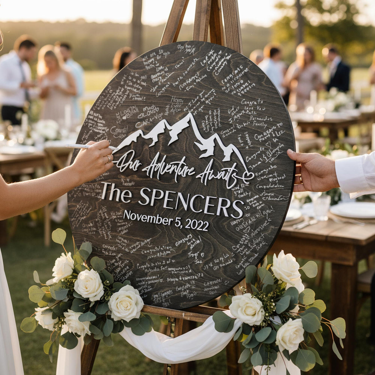 Black round mountain wedding guest book sign with raised white names and date on an easel, guests signing at an outdoor reception
