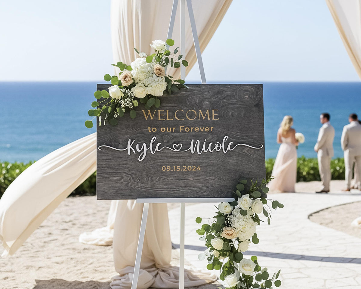 Grey wooden wedding welcome sign on a white easel at a beach ceremony, engraved Welcome to our Forever with the couple's names in raised white script