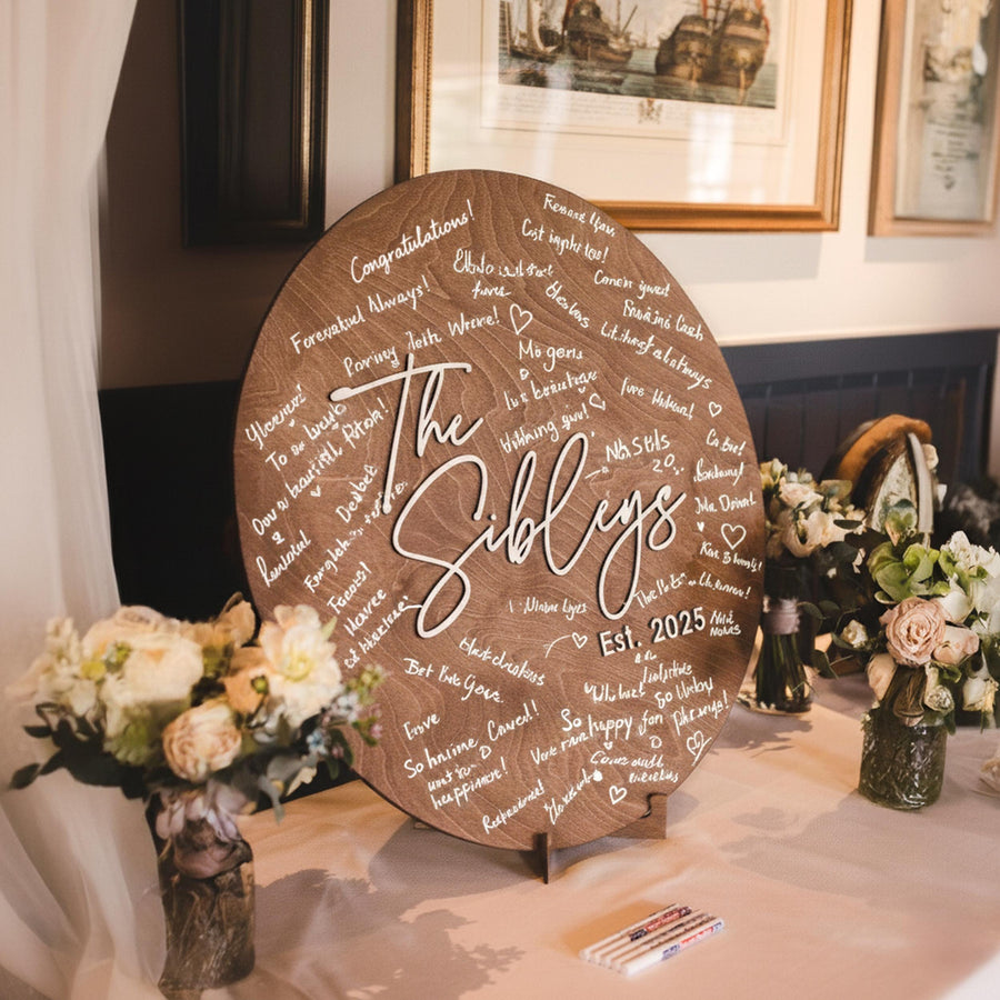 Warm brown round wooden guest book sign with a family name in white script on an easel beside a bouquet in a reception room.