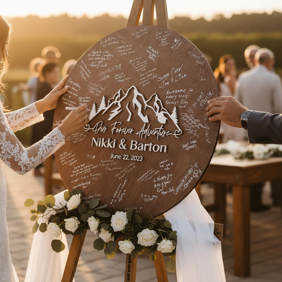 Dark brown mountain guestbook sign with raised white names and date held up on an easel with white flowers at an outdoor wedding.