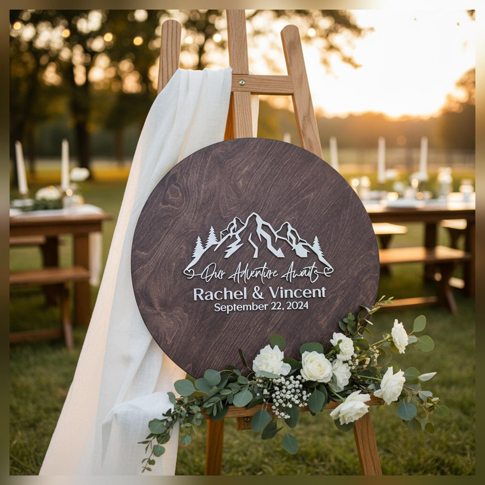 Dark brown mountain guestbook sign with names and date on an easel with a white drape and flowers at sunset.