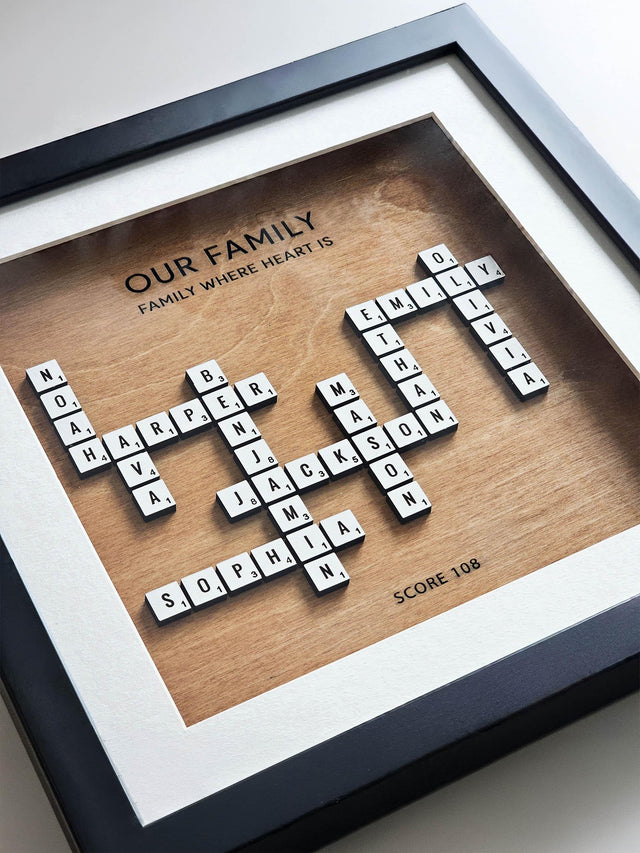 Close-up of the crossword family name frame: white letter tiles with point numbers on a hazelnut wood background, titled Our Family, inside a black frame.