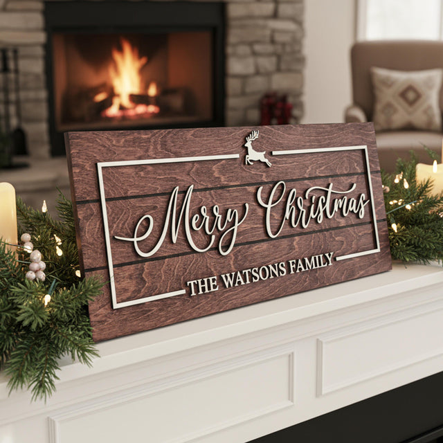 Personalized wooden Merry Christmas sign with reindeer icon and “The Watsons Family” text, displayed on a white mantel surrounded by candles, garlands, and a glowing fireplace in the background.