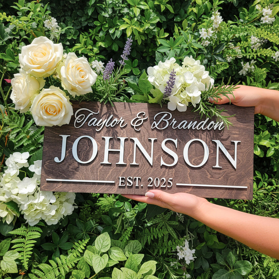 Family name sign reading Taylor & Brandon Johnson, Est. 2025 in raised white letters, held among white roses and greenery.