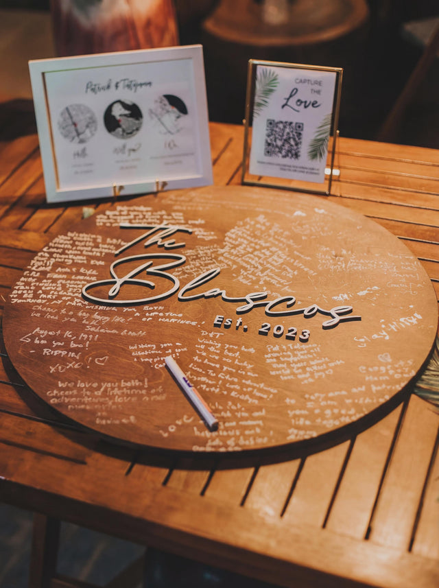 Warm brown round wooden guest book sign on a wooden table with a family name and year in raised lettering, covered in handwritten guest messages, with a white marker and two small framed signs behind it.