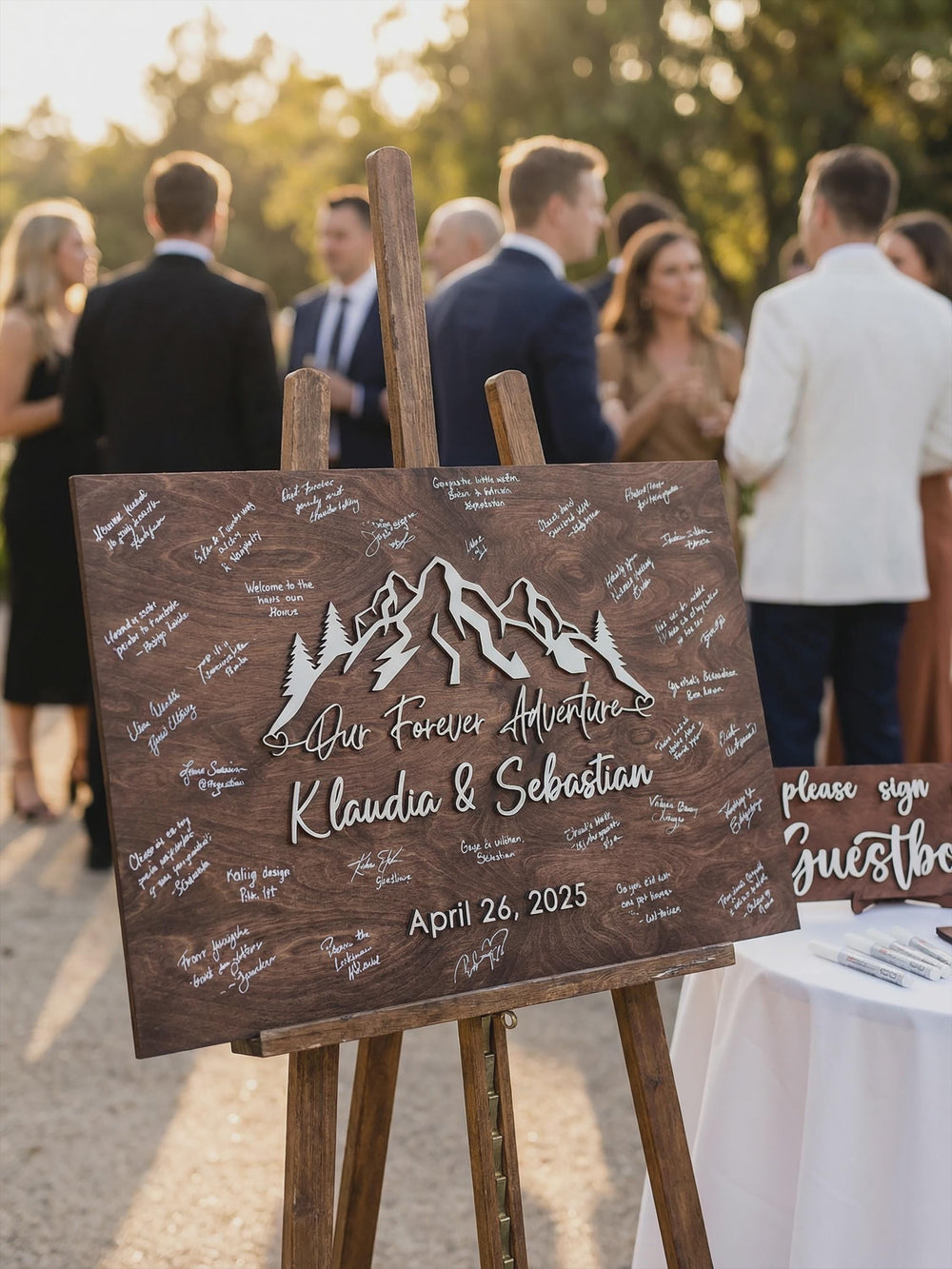 Forest mountain guest book sign on an easel at an outdoor reception, filled with signatures, beside the Please Sign Our Guestbook mini sign.