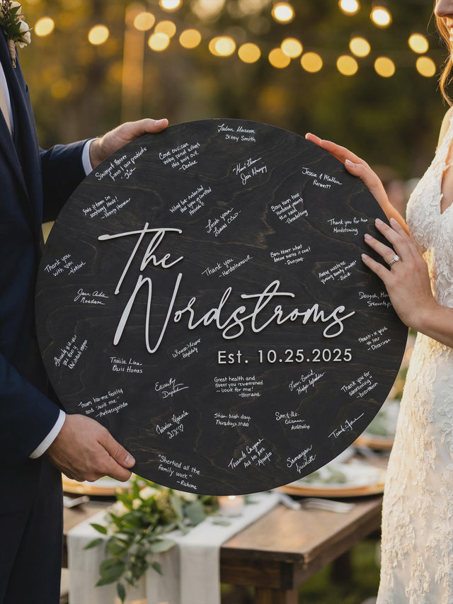 Black round wooden wedding guest book sign with a family name and date in raised white letters, held by the bride and groom at an evening reception.