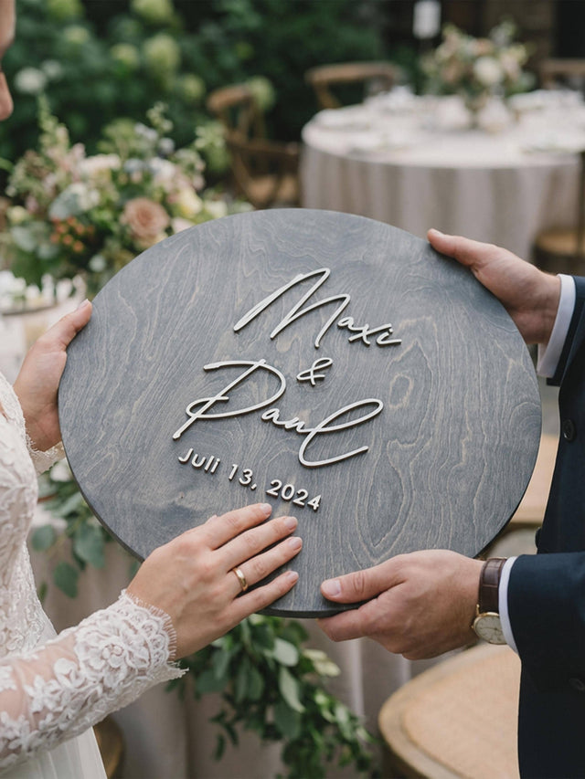 Grey round wooden wedding guest book sign with the couple's first names and date in raised white letters, held by the couple outdoors.