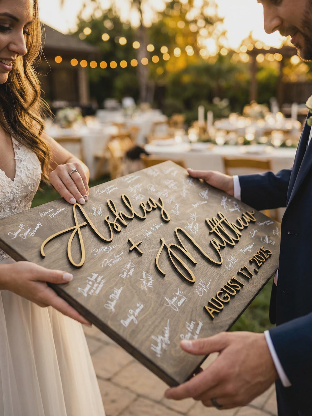 Bride and groom holding a personalized wooden wedding guest book alternative at an outdoor reception, featuring raised 3D names, a custom wedding date, and white handwritten guest signatures on a rustic dark stained wood board.