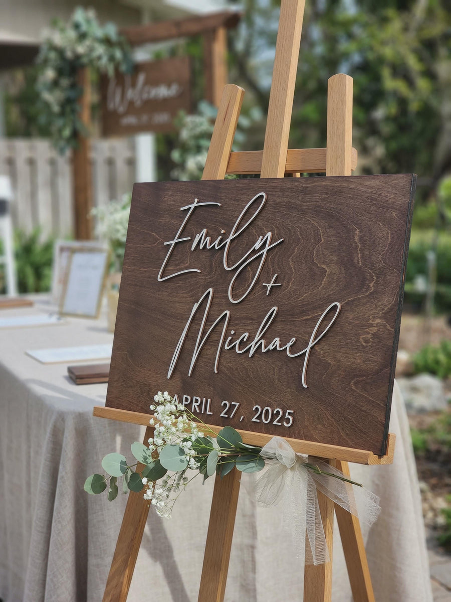 Personalized wedding guest book alternative on easel, rustic dark wood guestbook sign with 3D names and custom wedding date, displayed with greenery and tulle at outdoor wedding entrance.