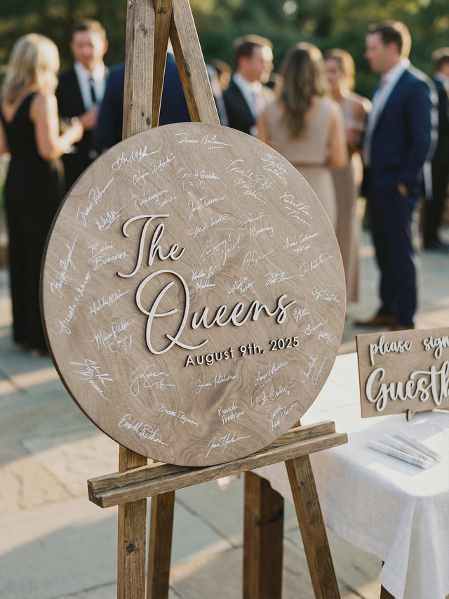 Round wooden wedding guest book sign on an easel at an outdoor ceremony, personalized with a family name and date in raised script lettering, with a small please-sign card beside it.