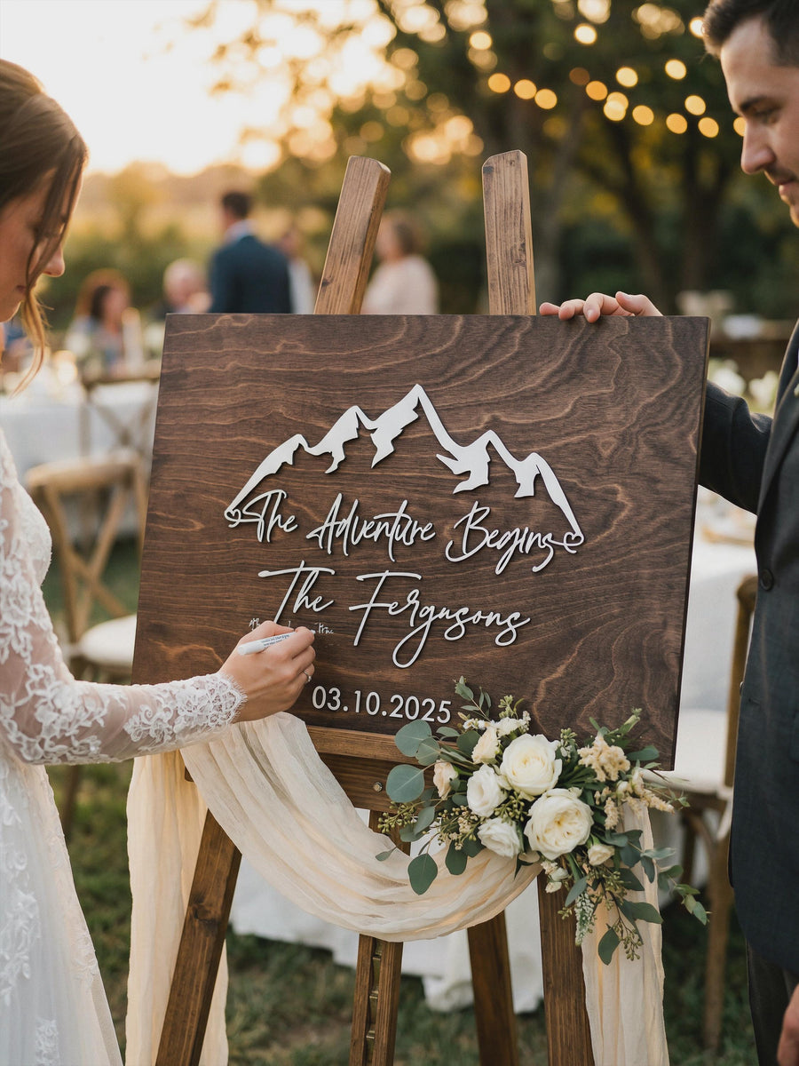 Bride and groom signing The Adventure Begins mountain guest book sign in Jacobean stain on an easel at an outdoor reception.