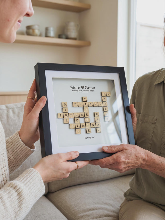 A younger and an older woman holding the black crossword family name frame between them on a sofa, natural wood letter tiles on a white background.