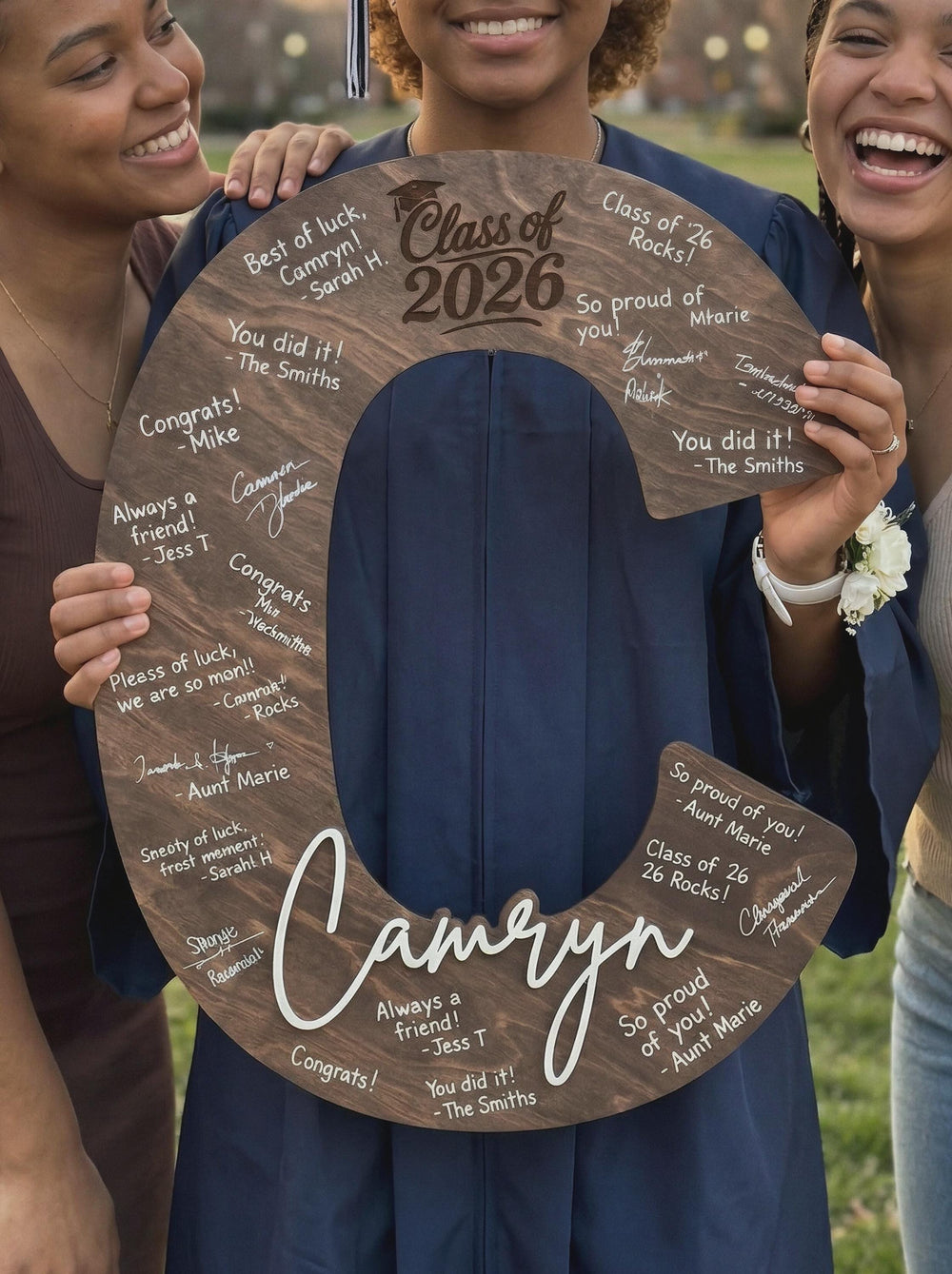 Two friends hugging a graduate as she holds a signed wooden letter C guest book.
