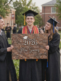 Graduate in a black cap and gown with two friends holding a dark wood Class of 2026 guest book sign, Max Simons raised in white script and messages written around it.