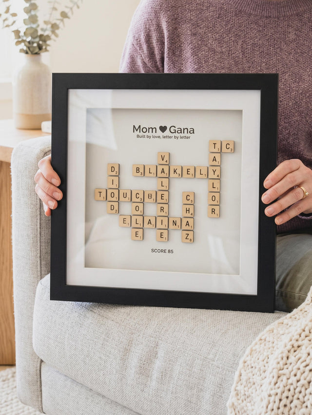 Crossword family name frame in a black frame with a white background, held on a sofa: natural wood letter tiles spell the family's names under the title Mom ♥ Gana.