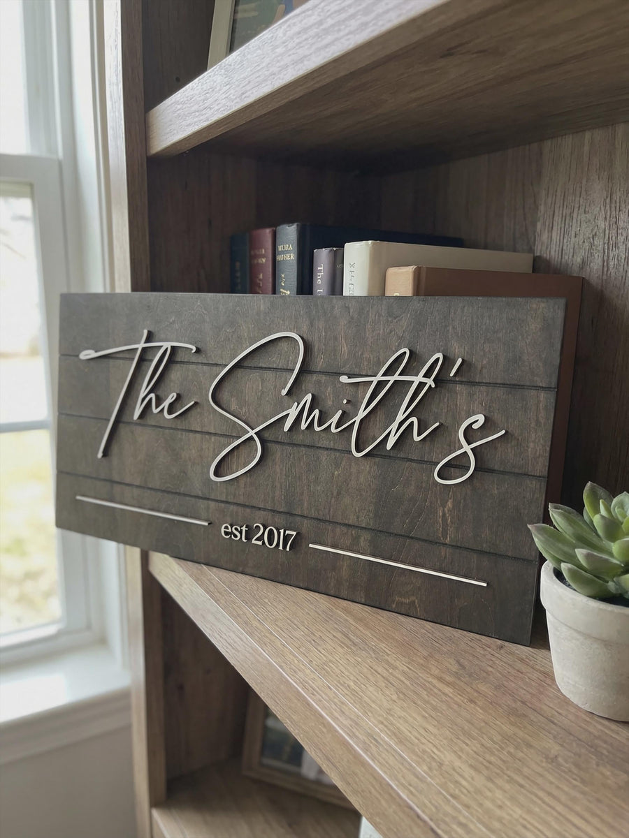 Last name sign reading The Smith's with est 2017 in raised white script, on a wooden shelf beside books and a small plant.