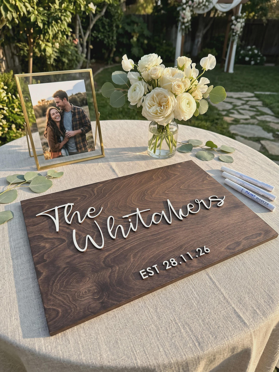 Rectangular Jacobean wooden guest book board with The Whitakers and EST 28.11.26 in raised white script, on a table with a photo frame, flowers and paint markers.