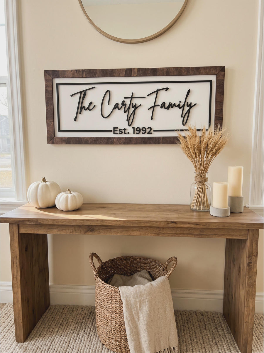 Framed family name sign in a Jacobean wood frame above an entryway console, The Carty Family and Est. 1992 in raised black script on a white backer.