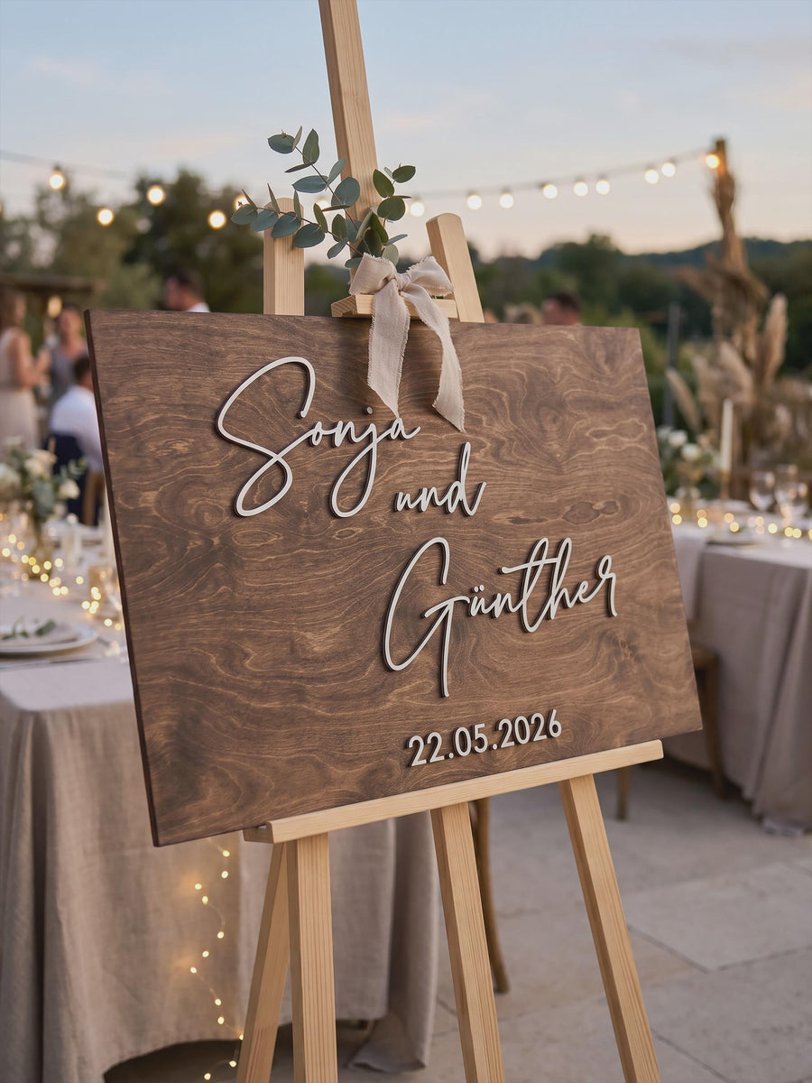 Rustic rectangular guest book sign with the couple's first names stacked in raised 3D script and the wedding date underneath, on an easel outdoors.