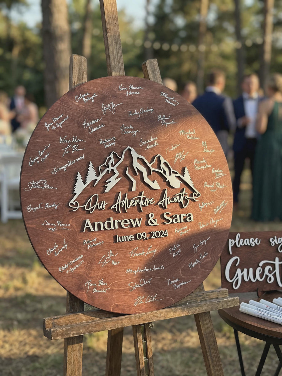 Brown mountain guestbook sign reading Our Adventure Awaits with the couple's names and date on an easel at an outdoor ceremony, beside the Please Sign mini sign.