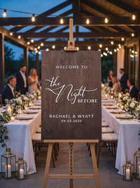 The Night Before welcome sign in a dark walnut finish with white raised letters, on an easel at an evening reception with string lights and set tables.