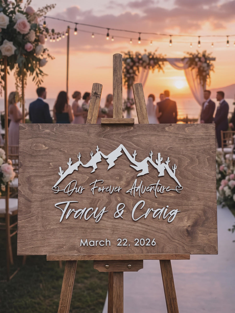 Desert cactus mountain guest book sign in Early American stain on an easel at a sunset beach wedding, with Our Forever Adventure, the couple's names and date in raised white letters.