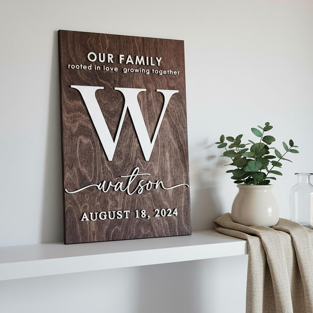 Portrait family monogram sign with Our Family, a large letter W, watson and the date August 18, 2024 in raised white letters, on a white shelf beside a plant.