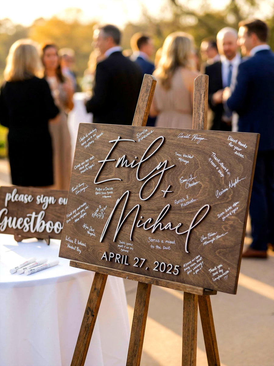 Wooden wedding guest book sign on an easel at an outdoor cocktail hour, with the couple's first names and wedding date in raised white script and guests' signatures, beside a Please Sign Our Guestbook mini sign.