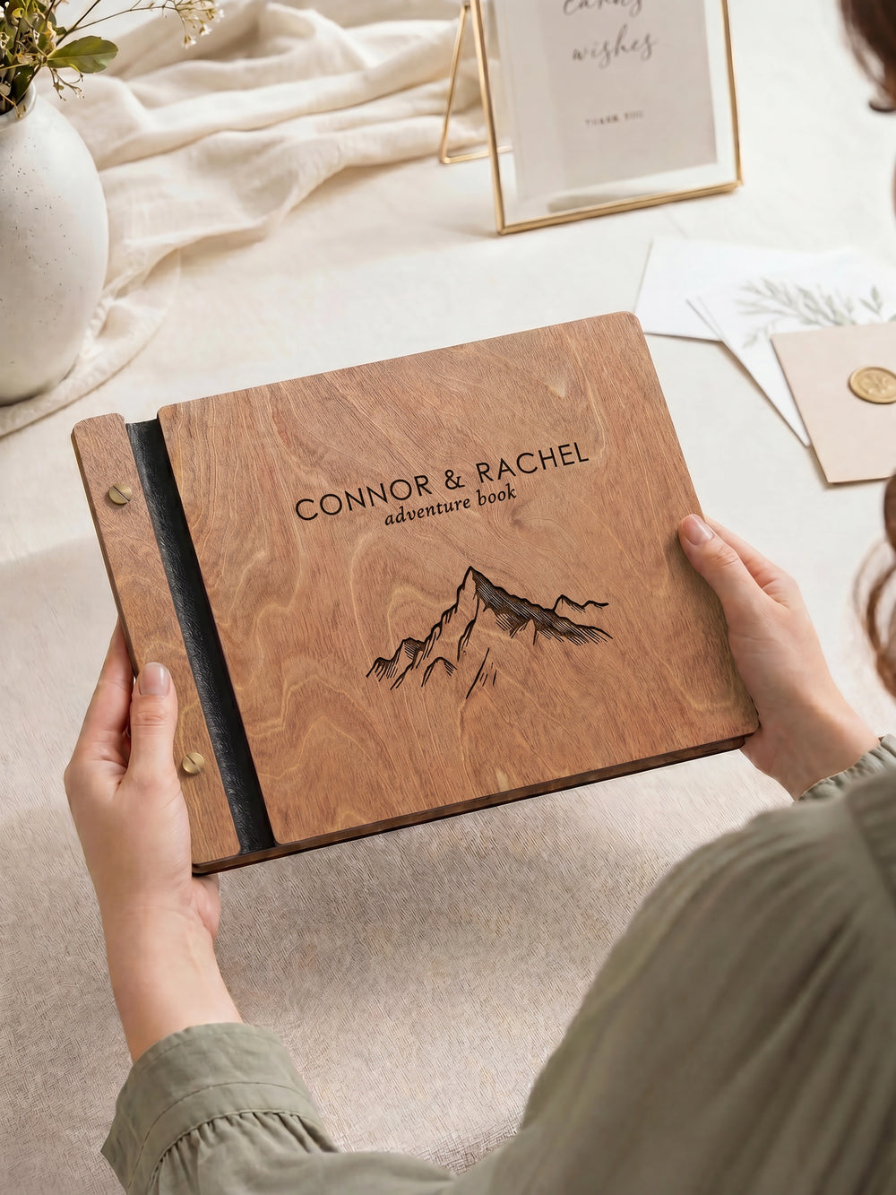 Hazelnut wood photo guest book engraved Connor & Rachel adventure book with a mountain drawing, held in two hands over a table with a card frame and photos