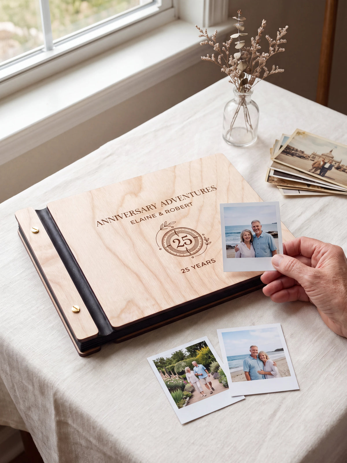 Natural wooden album with dark engraving of a circular 25-year anniversary emblem. A hand holds an instant photo beside the closed cover, with loose snapshots nearby.