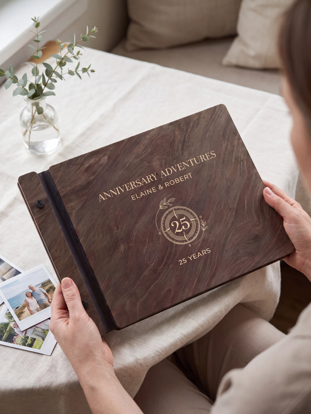Hands hold a dark brown Jacobean wooden album close to the camera. Cream engraving shows a circular 25-year anniversary emblem, personalized wording and the black binding strip.