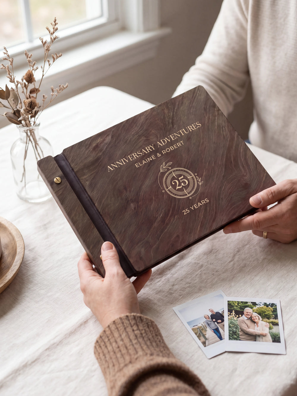 Two hands present a dark brown Jacobean wooden album, with the full cream-engraved circular 25-year anniversary emblem design and personalized cover wording facing the camera.