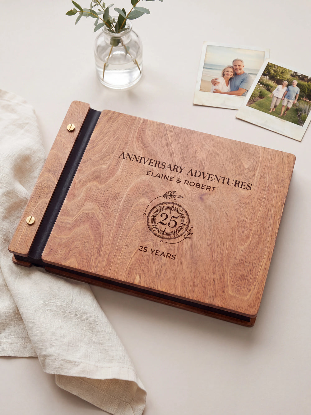 Hazelnut wooden album on a light tabletop with neutral linen. The full cover shows a circular 25-year anniversary emblem, personalized wording and the two-screw binding.