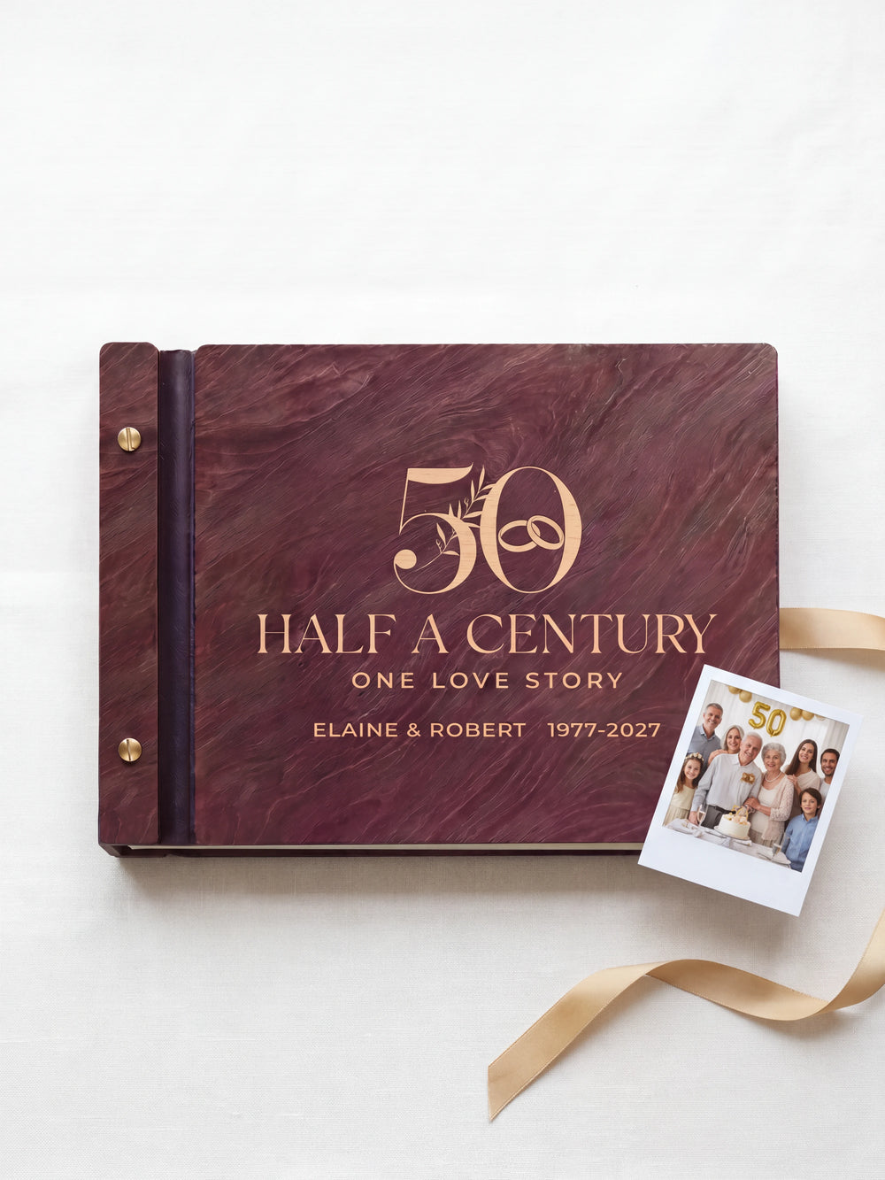 Jacobean wood 50th anniversary photo memory book lying flat on ivory linen, the cover engraved with “50”, “HALF A CENTURY, ONE LOVE STORY” and “ELAINE & ROBERT 1977-2027”; two gold-tone screws along the black vegan leather spine