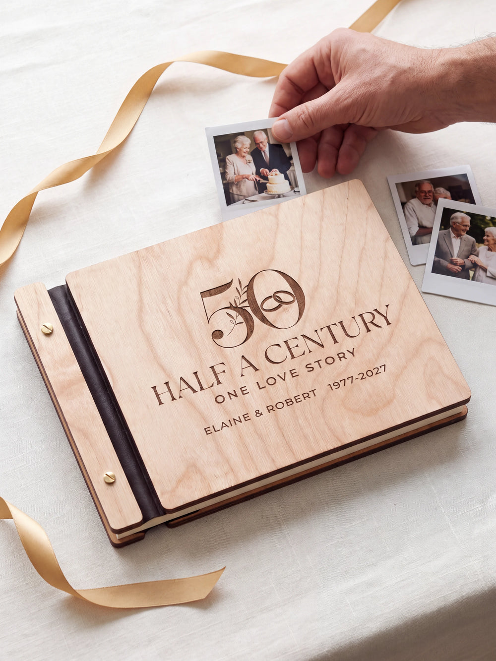 Natural wood 50th anniversary photo memory book on a light table while a hand picks an instant photo to add; the engraved cover reads “50”, “HALF A CENTURY, ONE LOVE STORY” and “ELAINE & ROBERT 1977-2027”