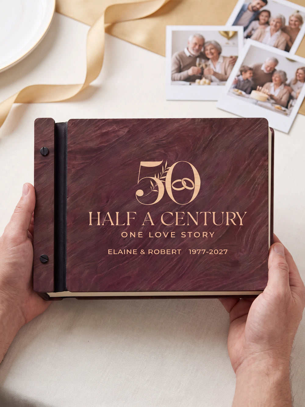 Two hands holding the Jacobean 50th anniversary photo memory book up to the camera, the cream engraving of a large number 50 with interlocking wedding rings and leaves and “50”, “HALF A CENTURY, ONE LOVE STORY” and “ELAINE & ROBERT 1977-2027” filling the cover