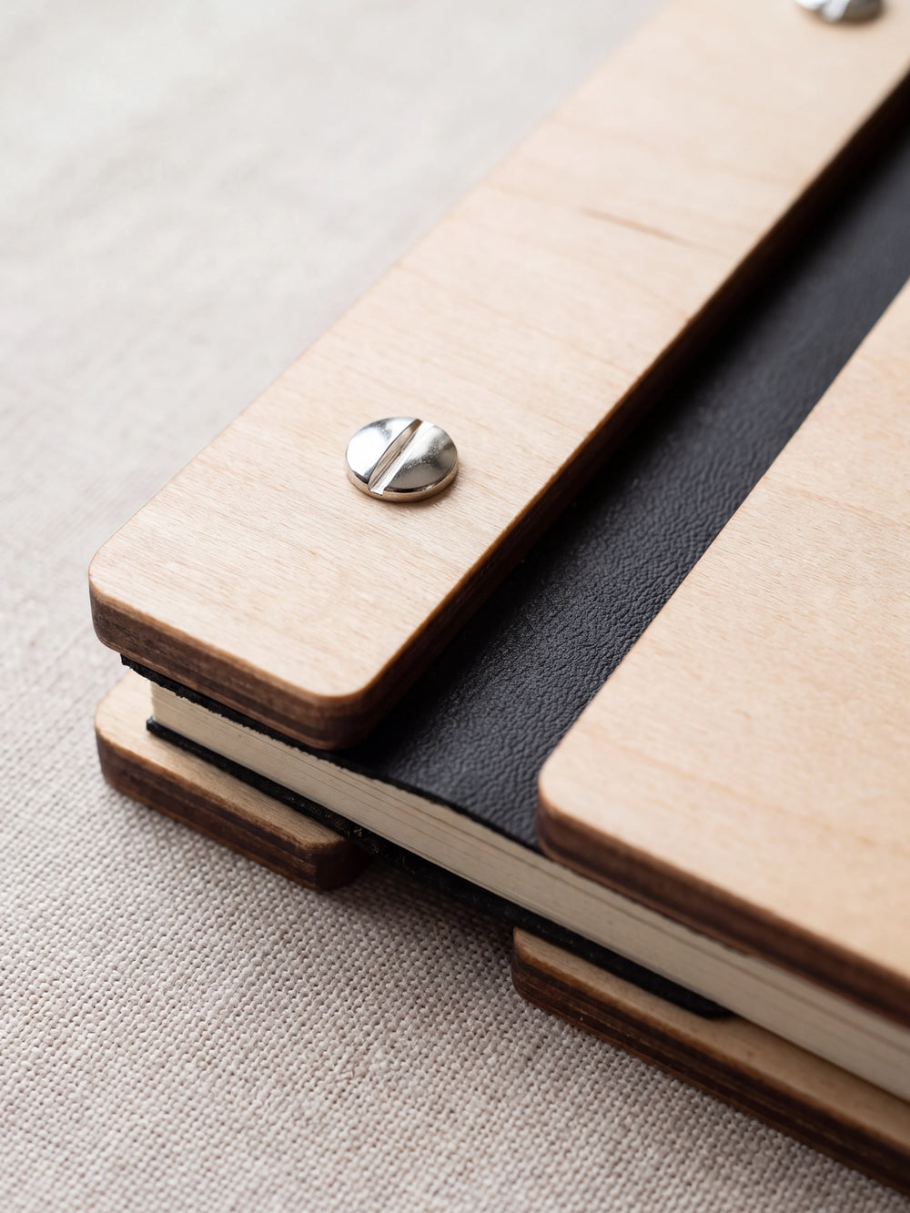 Close-up of a polished silver screw in the Natural wood strip beside the black vegan leather binding, the cream sheet stack visible below