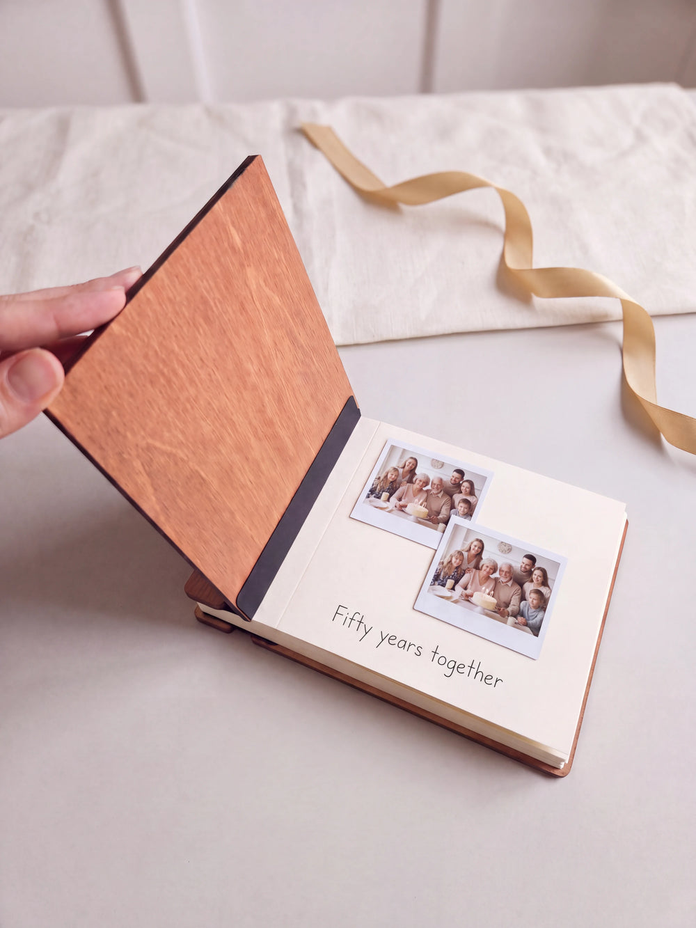 The Hazelnut 50th anniversary photo memory book open on a table with instant photos and a short handwritten note on a cream page; the black hinge strip is visible