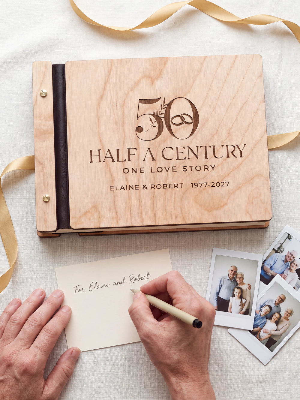 A hand writing a small gift card beside the closed Natural wood 50th anniversary photo memory book; the engraved cover and silver screws are visible