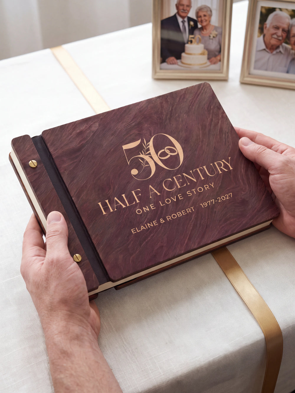 Two hands presenting the closed Jacobean 50th anniversary photo memory book with its cream engraving of a large number 50 with interlocking wedding rings and leaves, a single gold ribbon beside them