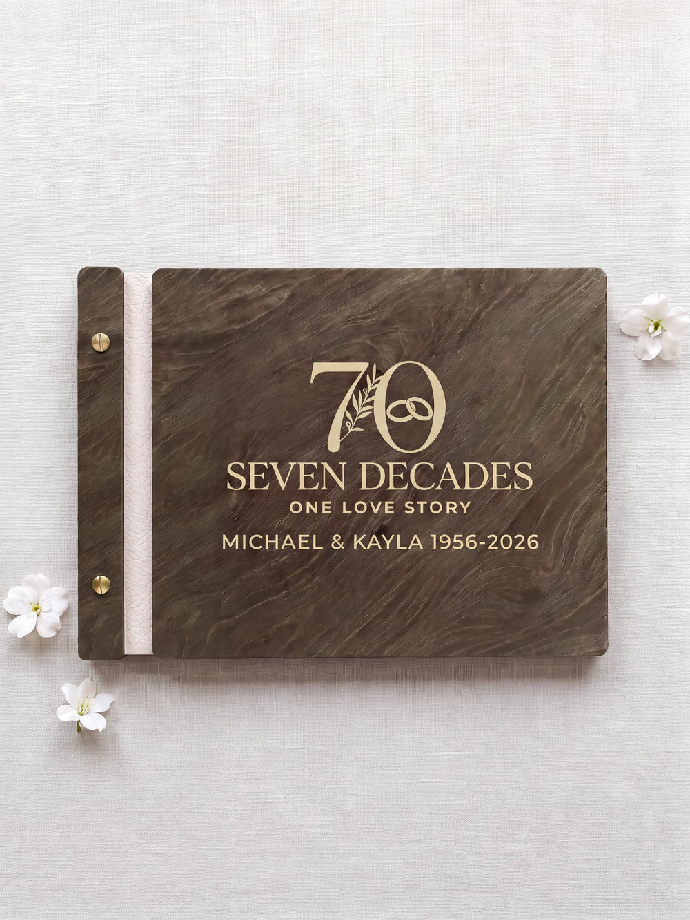 Jacobean wooden photo album with cream laser engraving of a large 70 with a botanical sprig and interlocking wedding rings. The closed book rests on pale linen with ivory binding and gold screws.