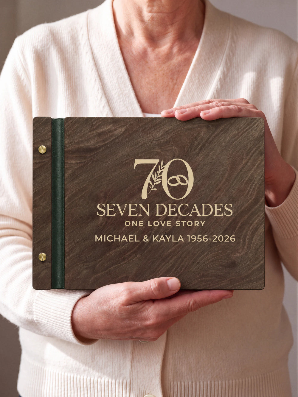 Jacobean wooden photo album with cream laser engraving of a large 70 with a botanical sprig and interlocking wedding rings. A person holds the cover toward the camera; forest green binding and gold screws are visible.