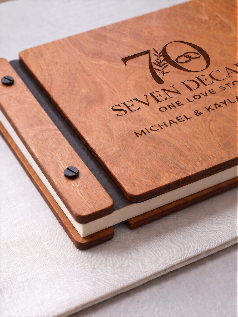 Hazelnut wooden photo album with dark brown laser engraving of a large 70 with a botanical sprig and interlocking wedding rings. A close cover view shows the engraved artwork, wood grain and screw binding.