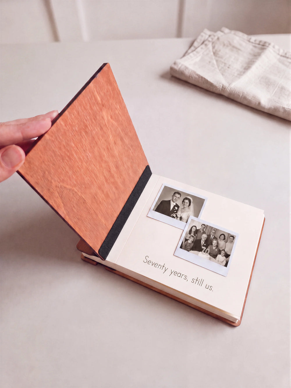 A hand lifts the Hazelnut album’s front wooden cover, revealing two photographs and a handwritten memory on a cream page. A thin black inner fabric strip lies on the wood.
