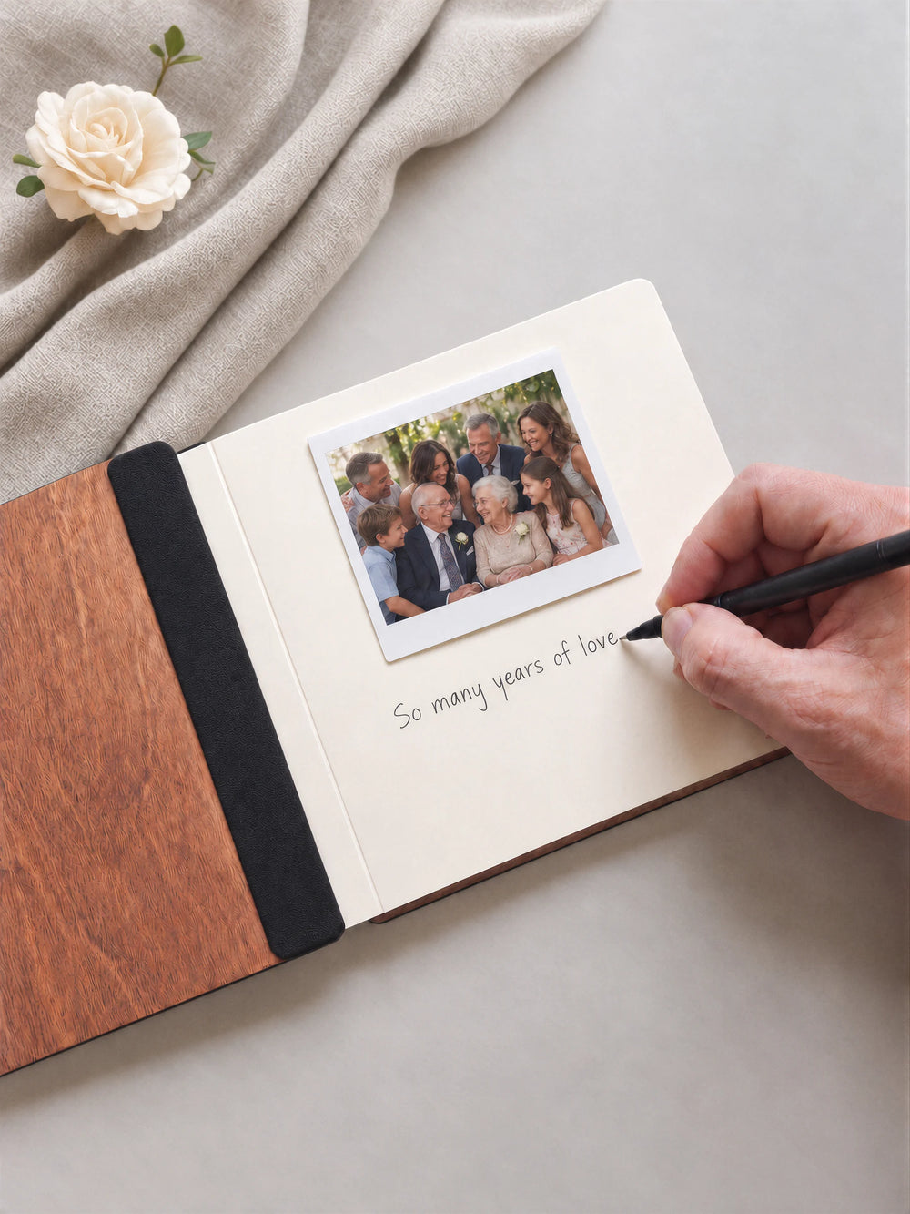 An open Hazelnut album with a photograph on a cream page. A hand writes beneath it; a flat black fabric strip runs along the inner wooden cover.