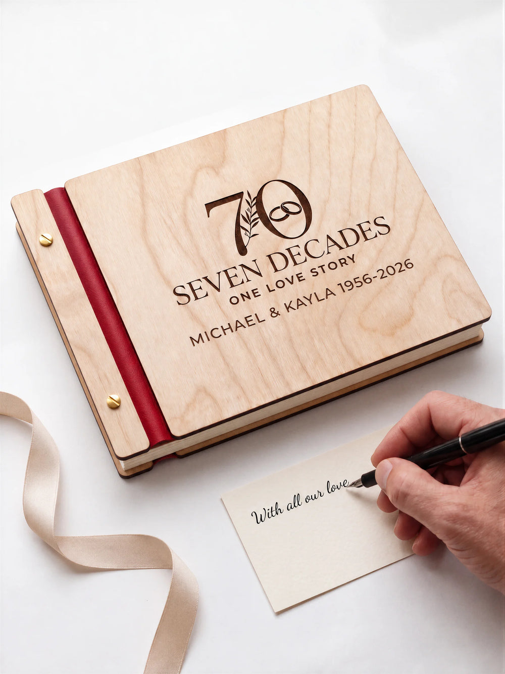 Natural wooden photo album with dark brown laser engraving of a large 70 with a botanical sprig and interlocking wedding rings. Cherry red binding and gold screws are visible while a hand writes on a separate gift card.
