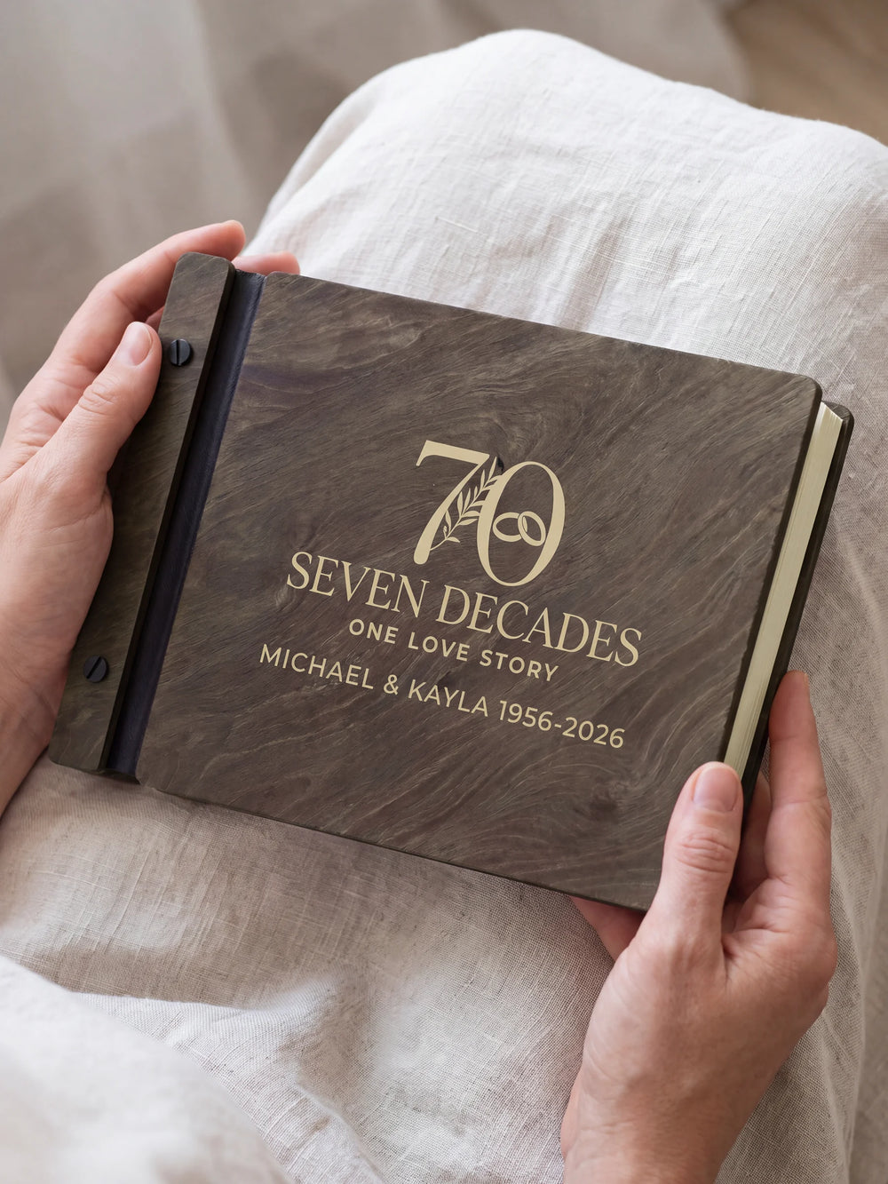 Jacobean wooden photo album with cream laser engraving of a large 70 with a botanical sprig and interlocking wedding rings. Hands hold the album close above pale fabric, with black binding and matte black screws.