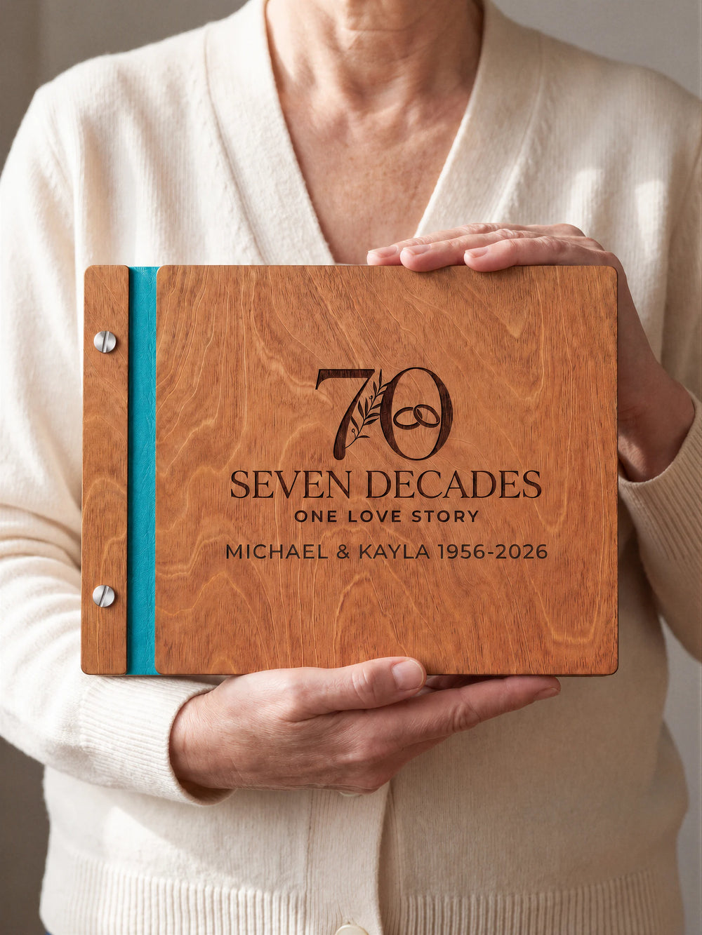 Hazelnut wooden photo album with dark brown laser engraving of a large 70 with a botanical sprig and interlocking wedding rings. A person holds the cover toward the camera, with turquoise binding and silver screws.