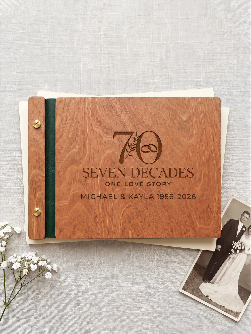 Hazelnut wooden photo album with dark brown laser engraving of a large 70 with a botanical sprig and interlocking wedding rings. The complete cover rests on linen beside an old photograph, with green binding and gold screws.